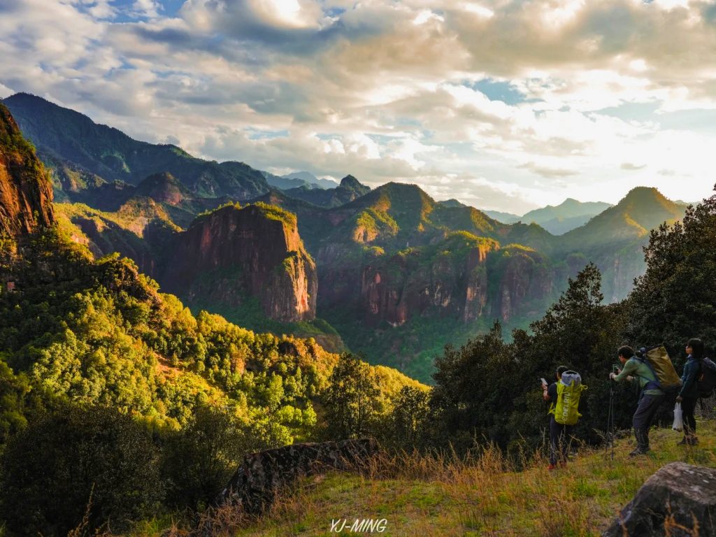 lijiang laojunshan limian danxia scenic area 麗江老君山黎明丹霞片區,夕陽柔光灑落在層疊紅色丹霞峰林,山谷覆蓋翠綠林木;數名登山客站在山巔眺望壯闊山景、拍攝風景,雲彩點綴天空,是老君山知名的丹霞地貌與戶外登山勝地。