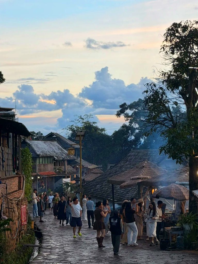 lijiang laojunshan liming village townscape 麗江老君山黎明鄉村寨,黃昏時分古樸石板街道人潮往來,木造傳統屋舍搭配茅草傘,炊煙裊裊。遠方天空佈滿大朵雲彩,林木環繞村落,是黎明丹霞景區內充滿在地風情的傈僳族鄉村市集景點。