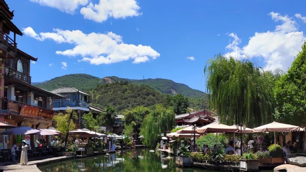 lijiang jiulongding scenic area town view 麗江九龍鼎風景區,清澈小河貫穿古鎮商街,兩側傳統建築與露天餐飲區,垂柳飄曳,遠方青山搭配湛藍天空,充滿滇西休閒風情。
