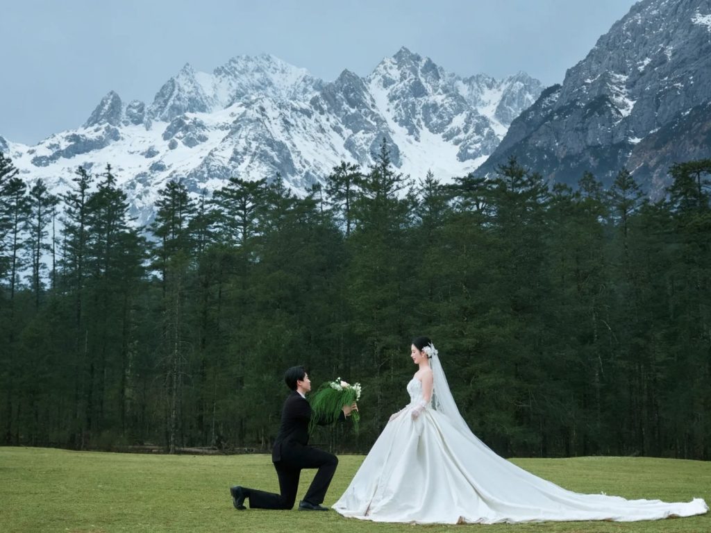 yulong mountain ganhaizi ranch wedding photo 玉龍雪山甘海子牧場婚紗照,新郎單膝下跪獻捧花,新娘身著白紗,背景為白雪覆蓋的山巒與蒼翠松林,浪漫戶外婚紗攝影。