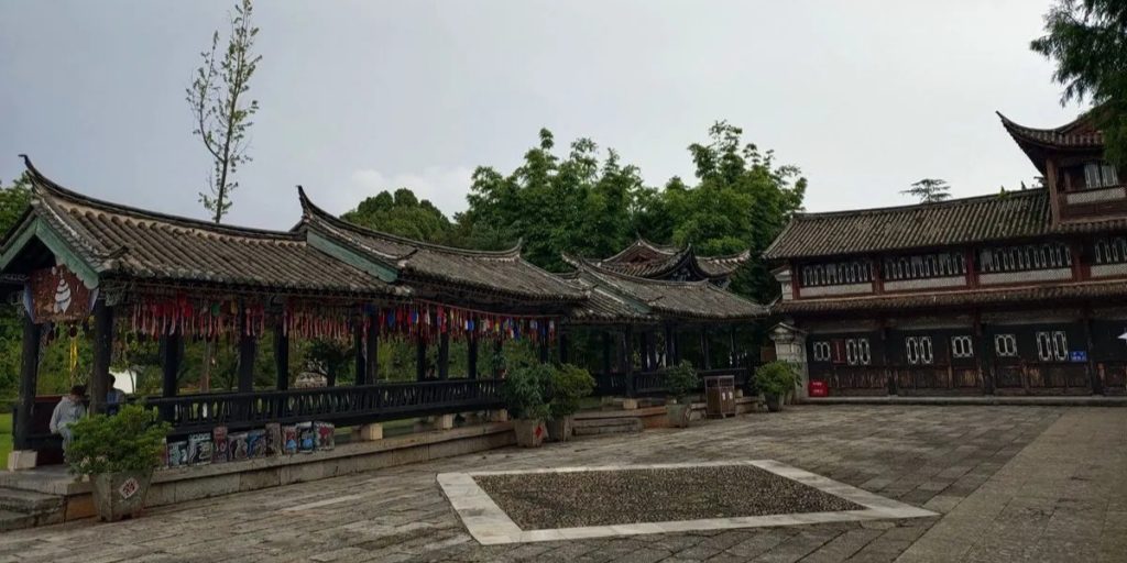 yunnan kunming nationality village naxi village 雲南昆明民族村納西族村寨,長廊式傳統木造建築搭配飛檐瓦頂,廊下懸掛多彩祈福吊牌,石砌廣場鋪設整潔,背景綠樹環繞,重現麗江納西族古建風格與村寨氛圍。