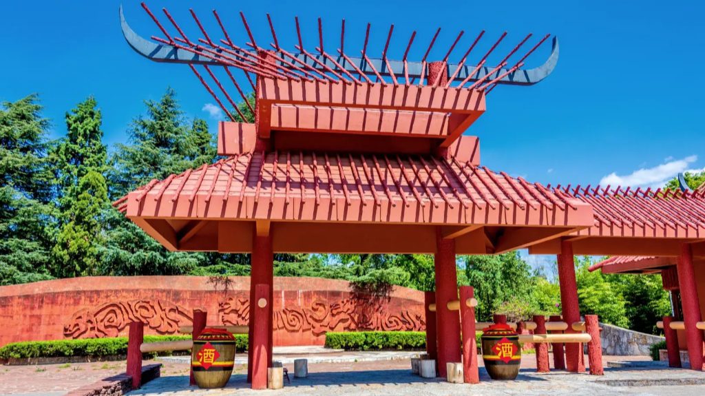 yunnan kunming nationality village yi ethnic village 雲南昆明民族村彝族村寨入口,紅色風格獨特的傳統牌樓,屋頂帶牛角造型裝飾,前方擺放印有酒字的大型酒缸,後方牆面刻有彝族圖騰浮雕,翠綠林木搭配湛藍晴空,呈現濃厚彝族文化特色。