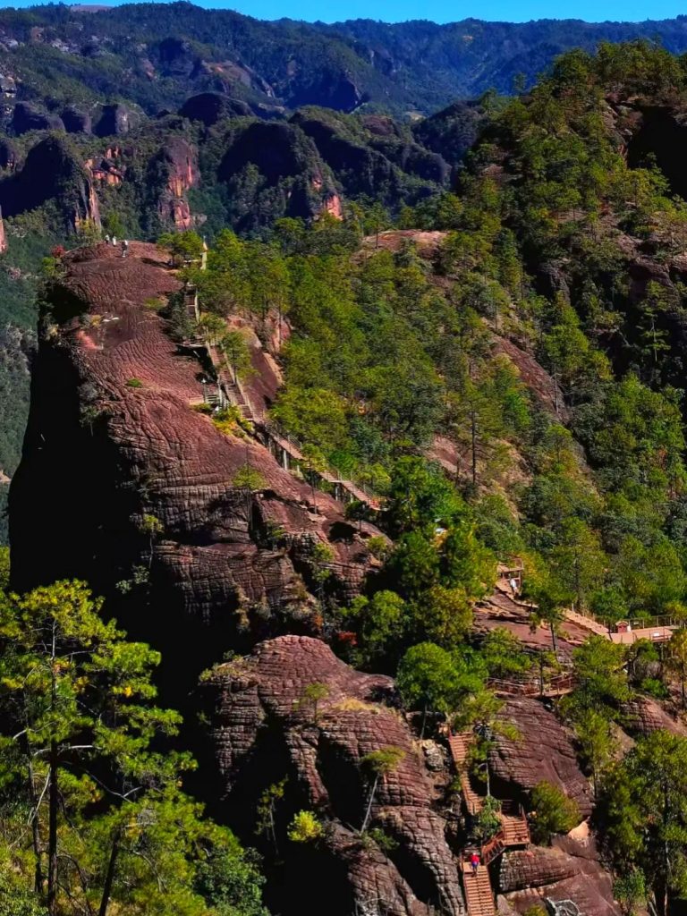 yunnan lijiang laojunshan limian qianguishan danxia landform 雲南麗江老君山黎明千鬼山丹霞地貌空拍,赤紅層理岩山聳立,岩壁鋪設木質登山步道與階梯,遊客漫步其間;山林翠綠環繞,連綿丹霞峰巒向遠方延伸,晴空之下呈現雄奇壯闊的紅岩地質景觀。