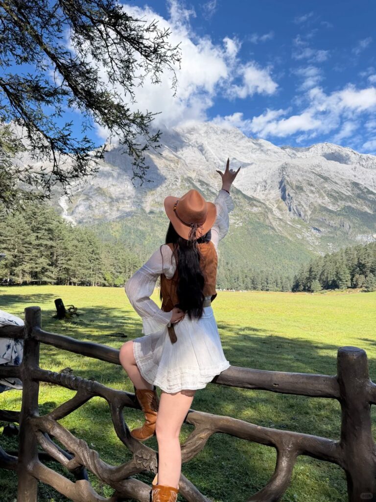 yunshanping travel portrait yulong mountain 麗江雲杉坪旅遊人像照,女子戴牛仔帽、穿波西米亞穿搭坐在木柵欄上,對著玉龍雪山比手勢,青綠草甸搭配藍天白雲。