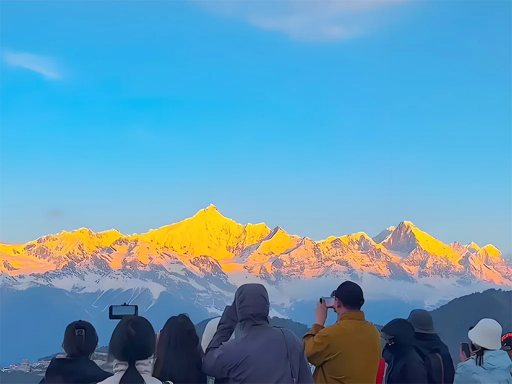 Diqing Meili Snow Mountain 1 迪慶梅里雪山日照金山壯麗景觀,清晨陽光灑落在連綿雪峰上,山頂染上金色光芒,前景遊客駐足拍照欣賞壯觀山景,展現迪慶梅里雪山著名的日照金山奇景與香格里拉高原自然魅力
