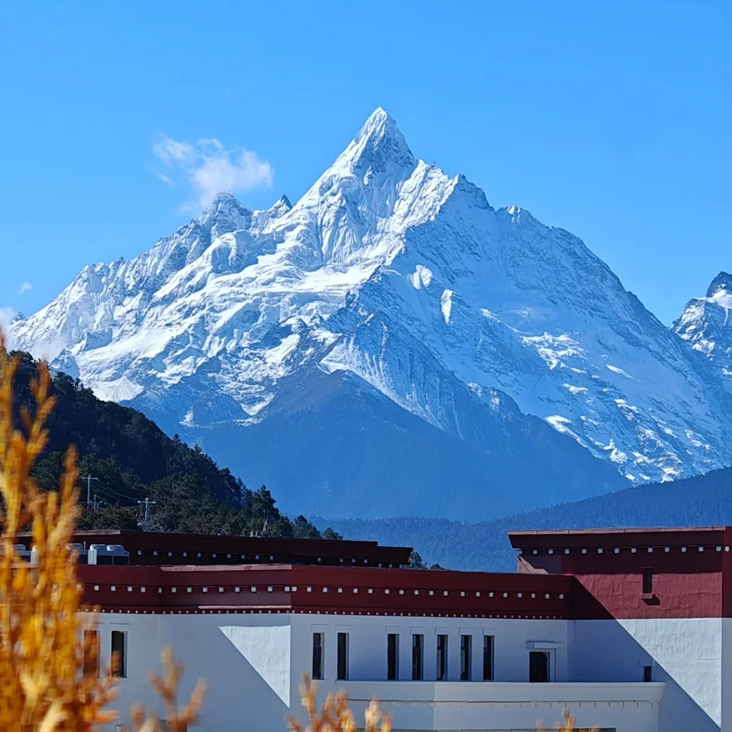 在 飛來寺 遠眺，雪山在陽光照耀下潔白耀眼，山峰巍峨聳立，前景建築與遠山形成層次分明的壯麗景觀。