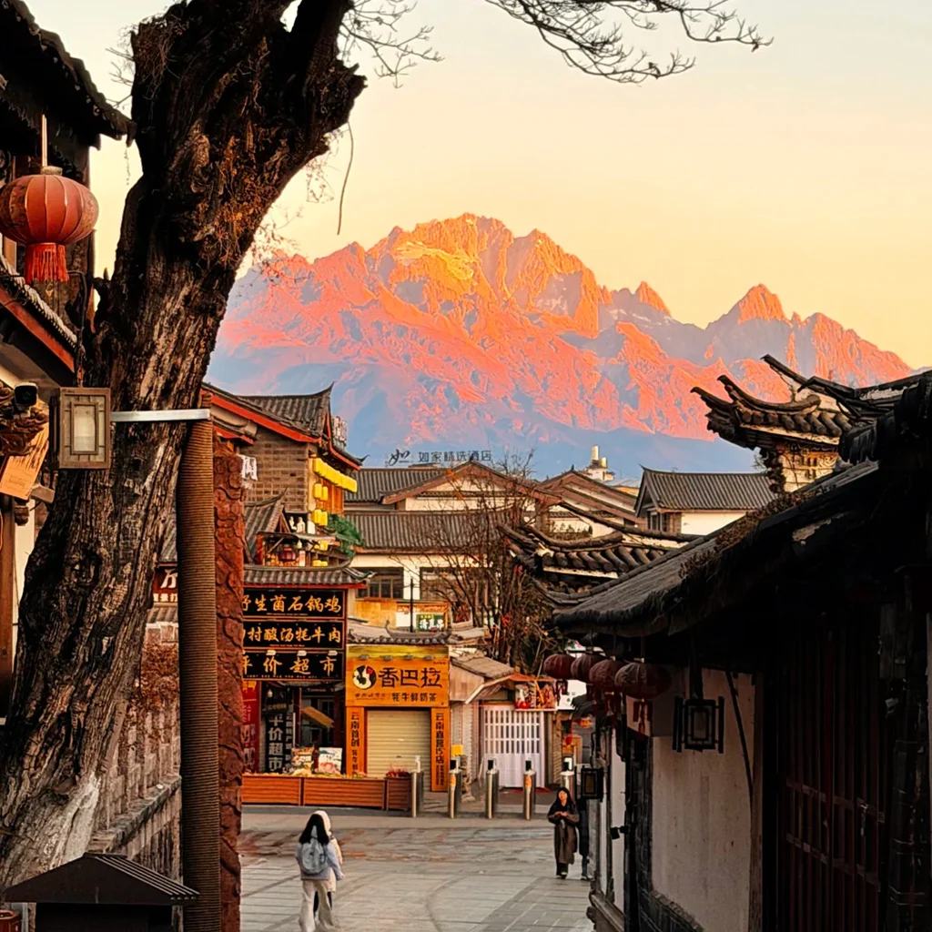 清晨的 麗江古城，傳統瓦屋、靜謐石板巷道，遠方是被晨光染金的玉龍雪山。