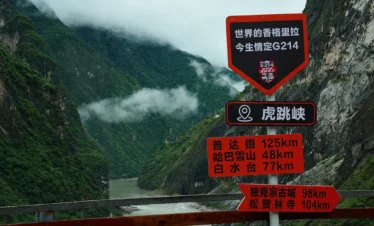 虎跳峽 G214 國道上的網紅打卡路牌，「世界的香格里拉，今生情定 G214」字樣格外醒目，背後是洶湧的金沙江與雲霧繚繞的峽谷。