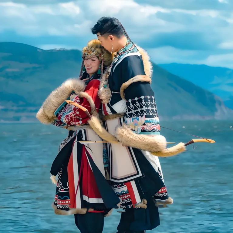 在香格里拉湖畔，一對身穿藏族傳統服飾的男女站在湖邊合影。服裝以鮮豔色彩與毛皮裝飾為特色，展現濃厚民族風情。背景是清澈湖水與遠方群山，藍天雲層映襯高原風景，呈現香格里拉獨特的人文文化與自然景觀。