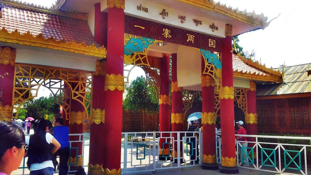 One village two countries 一寨兩國景區入口建築,紅色立柱與金色裝飾呈現東南亞民族風格,上方牌匾寫著「一寨兩國」,遊客在入口處參觀拍照,展現中緬邊境一寨兩國獨特的地理與文化景觀。