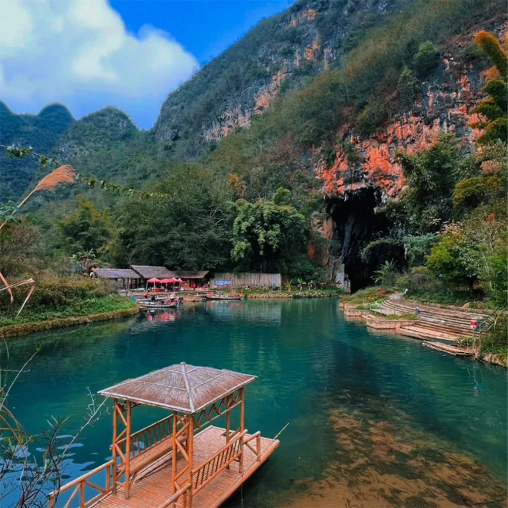 位於 坝美溶洞 的湖泊與溶洞入口景觀。清澈湖水倒映群山與森林，岸邊設有竹木亭與步道，遠處紅色岩壁下的巨大溶洞口通向山體內部。