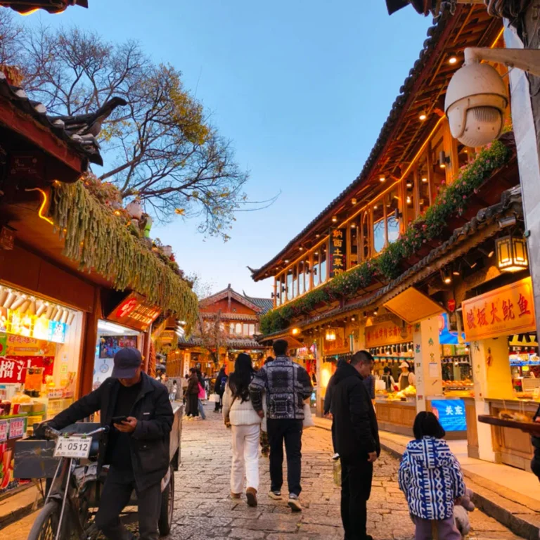 Lijiang Old Town 雲南著名歷史景區 麗江古城 的傍晚街景。石板路兩側是傳統木質建築與商店,屋簷掛著燈飾與招牌。遊客與當地居民在古街上漫步購物,街道燈光溫暖,營造出熱鬧又富有文化氣息的古鎮氛圍,展現麗江古城獨特的旅遊魅力