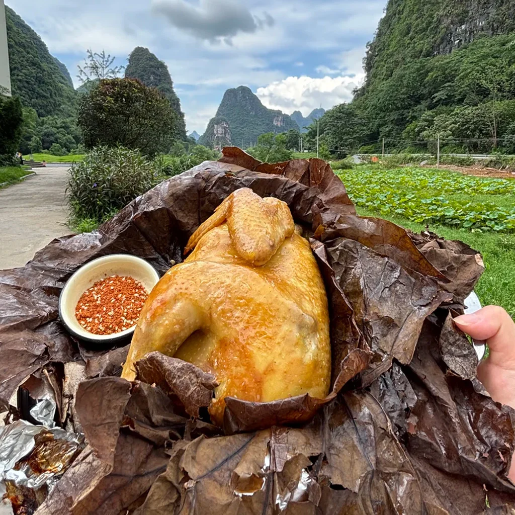 雲南文山普者黑的道地荷葉雞。多汁的整隻雞以香氣撲鼻的荷葉包裹，搭配辣椒蘸料，背景是喀斯特山景。