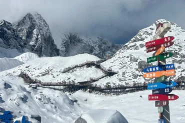 Montagna innevata del Drago di Giada 1 Punto panoramico a 4506 sul Monte Yulong a Lijiang: le cime innevate e frastagliate sono ricoperte da una coltre di neve candida; una passerella di legno si snoda tra le montagne innevate, mentre le cime sono avvolte da nebbia e nuvole; sono presenti cartelli che indicano la distanza dalle città cinesi e straniere.