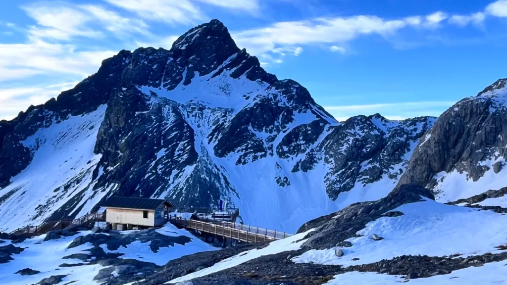 Jade Dragon Snow Mountain 3 雲南麗江玉龍雪山,雄偉積雪山峰拔地而起,山腳有木棧道與山中小屋,背景是澄澈藍天。