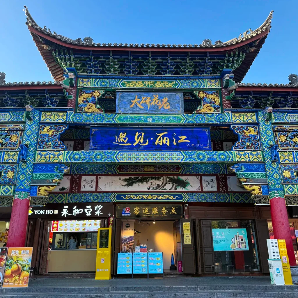 Lijiang Ancient Town — Dayan Flower Lane 雲南麗江古城大研花巷,雕梁畫棟的傳統中式牌樓,「遇見・麗江」字樣,下方是在地店家,背景是澄澈藍天。