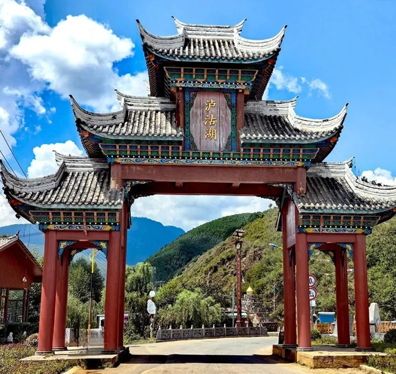 瀘沽湖景區入口的傳統中式牌樓矗立在道路上方，紅色立柱與多層飛簷屋頂展現典雅建築風格。牌樓中央寫著「瀘沽湖」三字，背後是綠色山巒與藍天白雲，呈現通往瀘沽湖自然風景區的地標入口與濃厚地方特色。