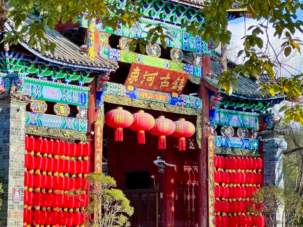 Shuhe Ancient Town 3 麗江束河古鎮入口,雕梁畫棟的傳統中式牌樓,掛滿紅燈籠與「束河古鎮」牌匾,背景是澄澈藍天。