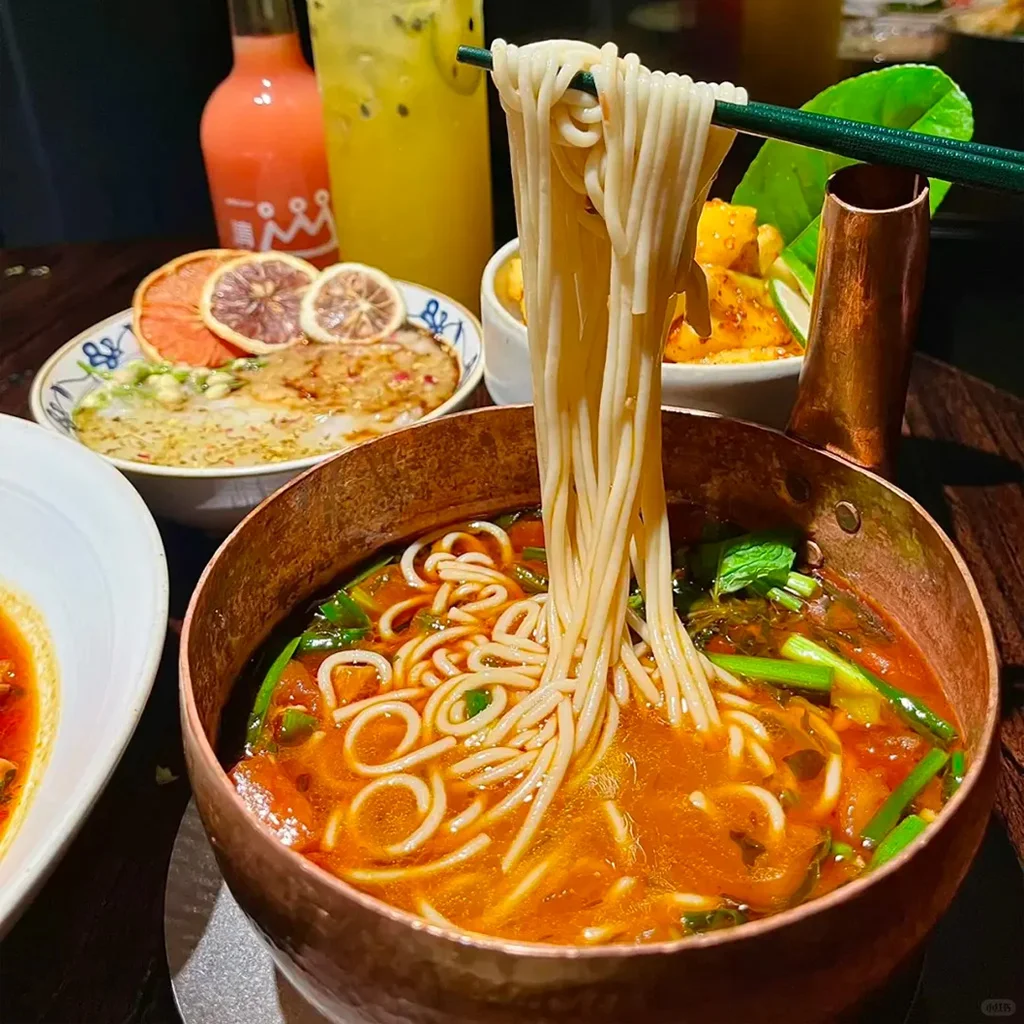 Yunnan Small Pot Rice Noodles 道地雲南小鍋米線盛放在手工錘紋銅鍋中。香辣紅湯、新鮮香草,以筷子夾起的米線,搭配雲南風味小菜與飲品。