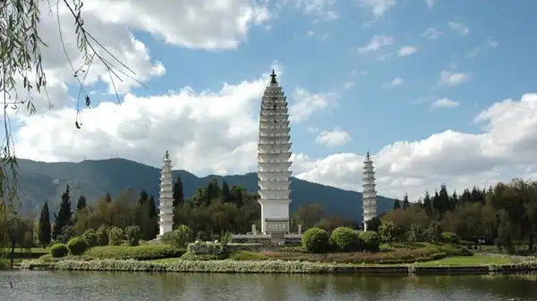 yunnan kunming nationality village bai village three pagodas 雲南昆明民族村白族村寨三塔景觀,三座白色佛塔臨湖而立,主塔高聳、兩座副塔對稱,湖畔綠草樹木環繞,遠處山巒與白雲相映,仿大理崇聖寺三塔,還原白族經典古建風貌。