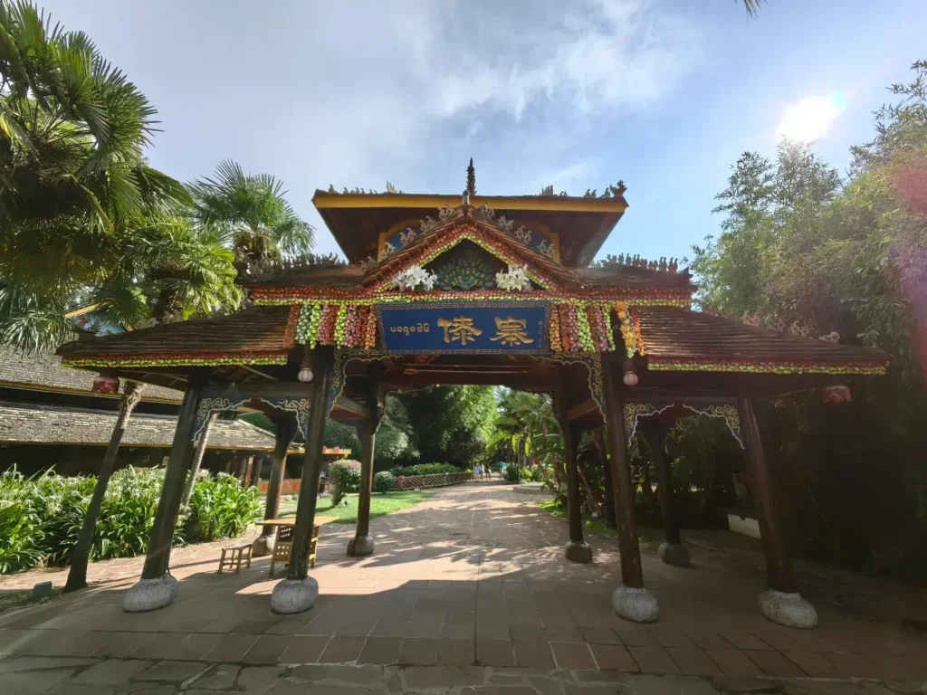 yunnan kunming nationality village dai ethnic village 雲南昆明民族村傣族村寨入口牌樓,傣式重檐木構建築,屋頂飾孔雀雕花與鮮花串,牌匾寫有傣文與漢字「傣寨」,四周棕櫚等熱帶植栽環繞,石磚步道延伸入村寨,充滿濃厚傣族風情。