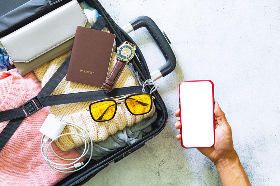 yunnan travel essentials luggage mobile prep 敞開的行李箱內裝有毛衣、護照、手錶、墨鏡與充電線,右側手持一部空白螢幕的手機